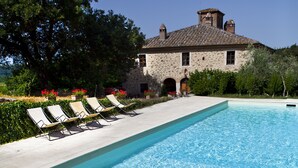 Une piscine rectangulaire à l’eau d’un bleu limpide, entourée d’une terrasse en pierre. Des chaises longues sont alignées le long du bord de la piscine, et un bâtiment en pierre avec un toit en tuiles est visible au fond.