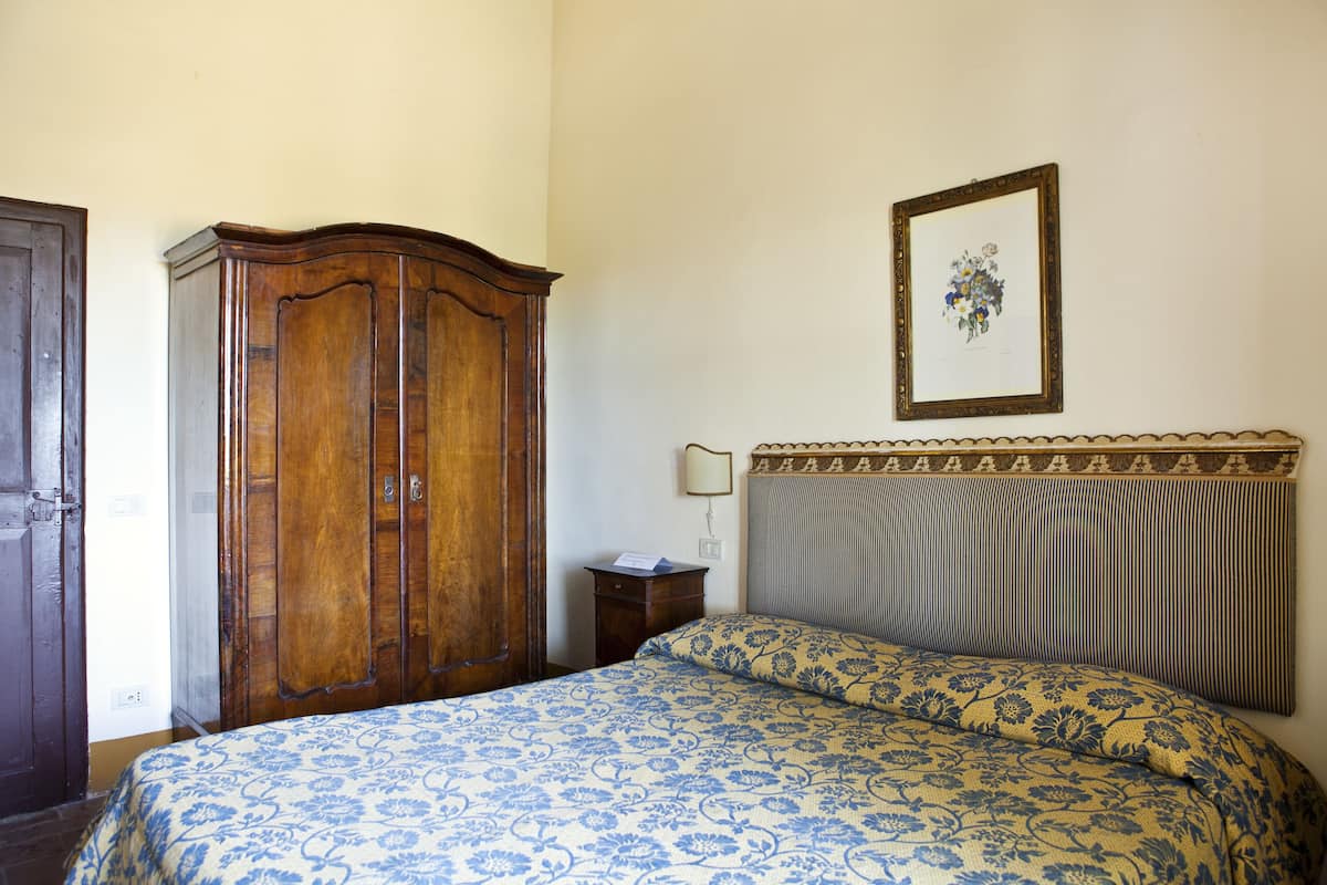 Ein Schlafzimmer mit Holzschrank, einem Bett mit blumigem Überwurf und einem Nachttisch mit Lampe. An der Wand hängt ein gerahmtes Bild.