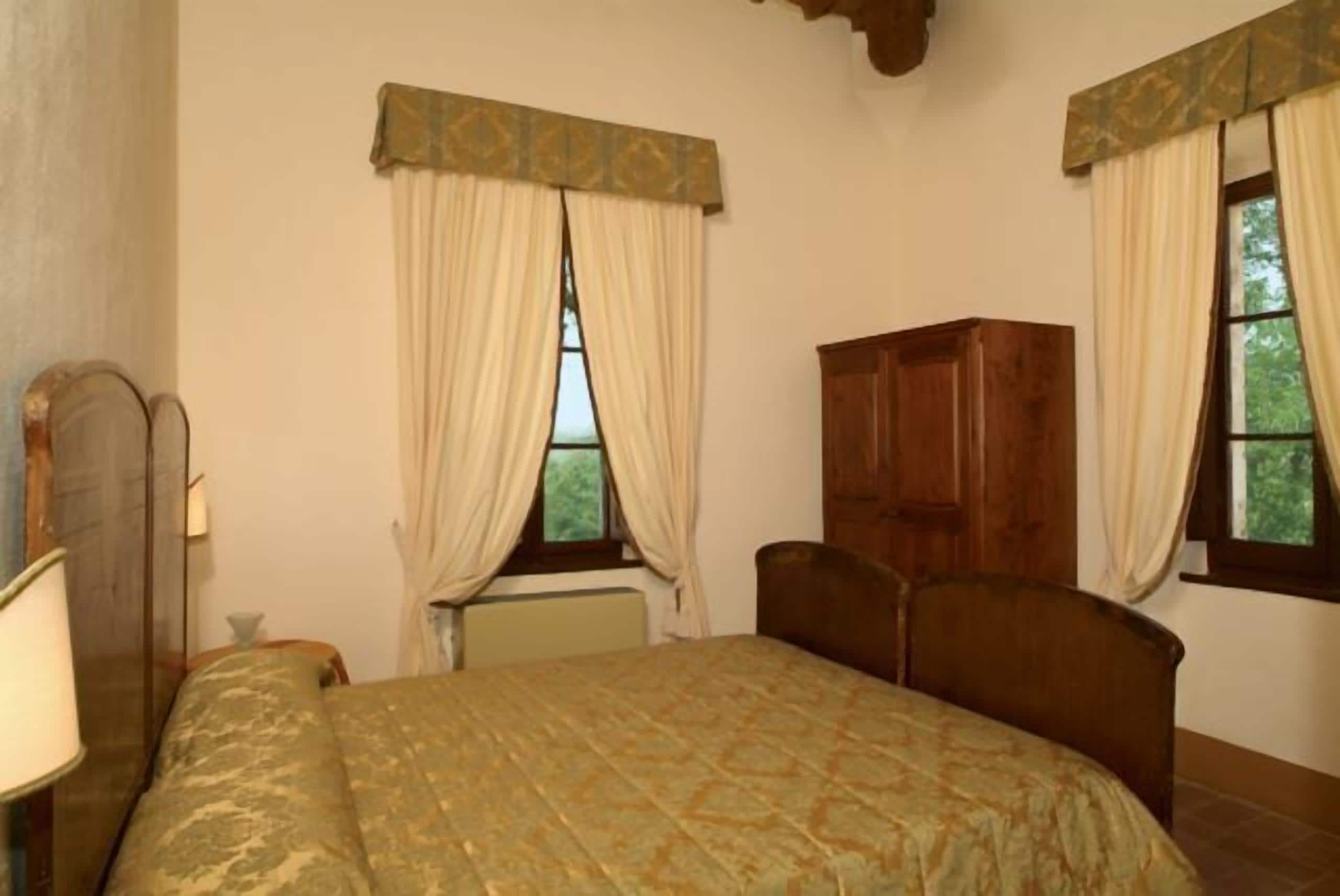 A bedroom with a bed, a wooden wardrobe, and two windows with curtains.