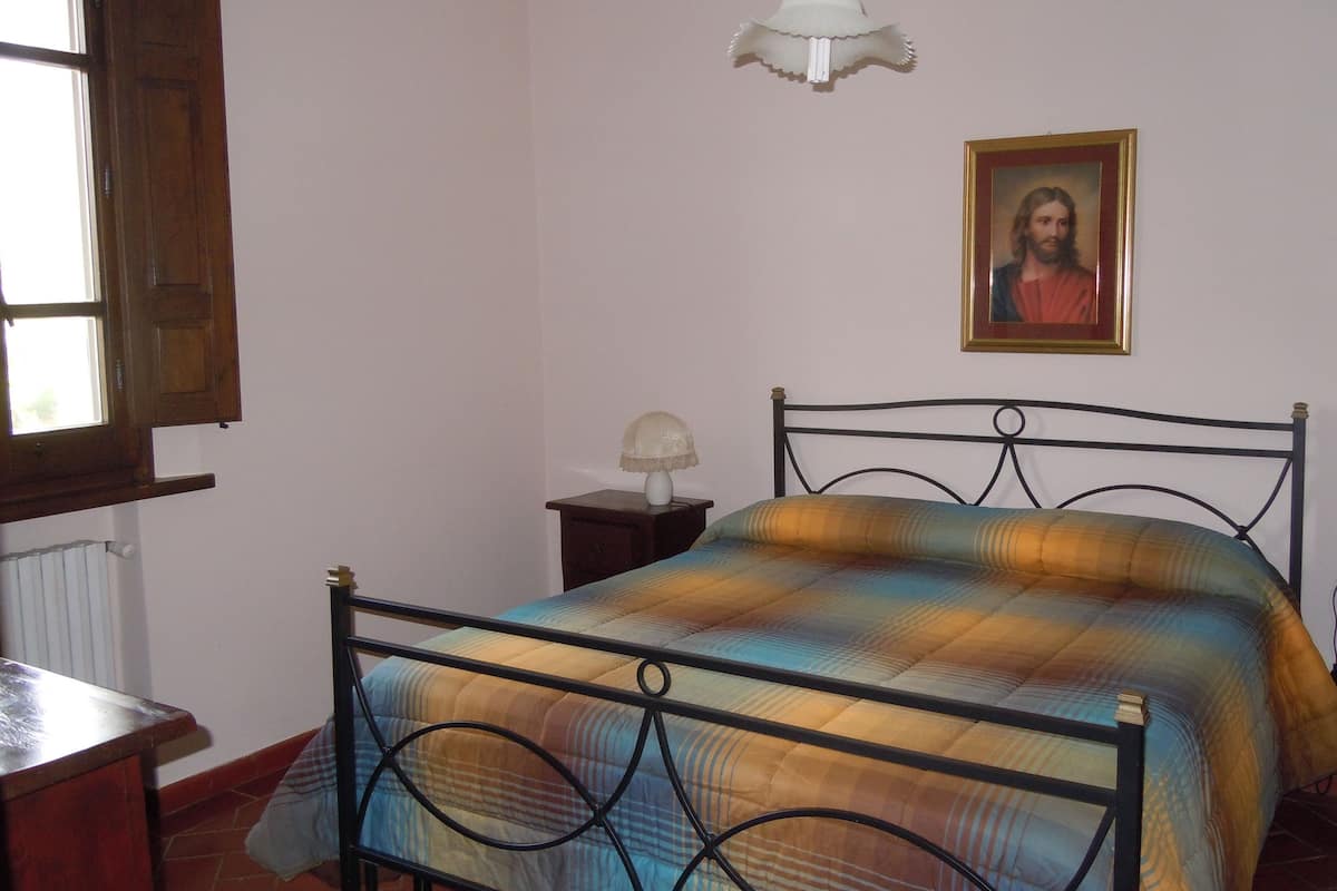 A bedroom with a metal bed frame, a bedside table with a lamp, a window with wooden shutters, and a framed picture on the wall.