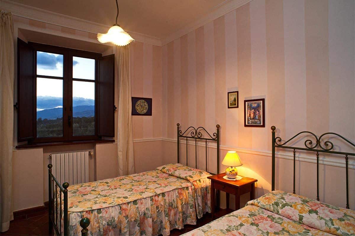 A bedroom with two single beds, a window with a view, a radiator, and a bedside table with a lamp.