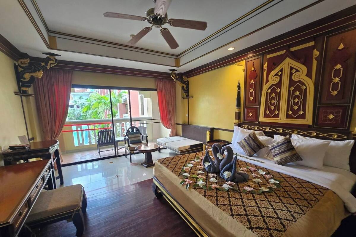 A hotel room with a bed, a desk, a chair, a balcony with a view of greenery, and a ceiling fan.