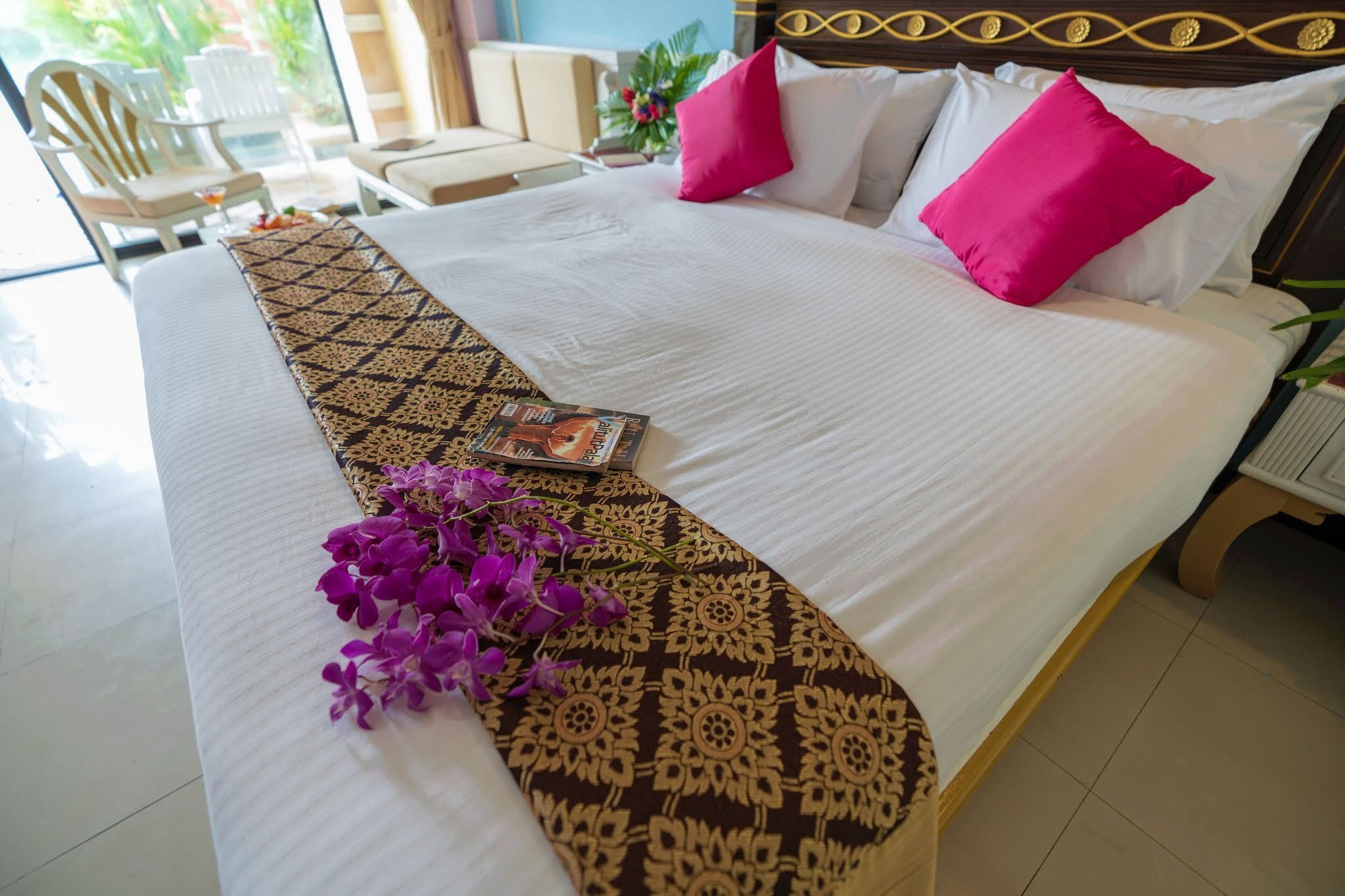 A neatly made bed with a patterned bedspread, two bright pink pillows, and a bouquet of purple flowers.