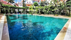 Una piscina azul cristalina con borde de azulejos, rodeada de exuberante vegetación y un edificio tradicional con arquitectura ornamentada al fondo.