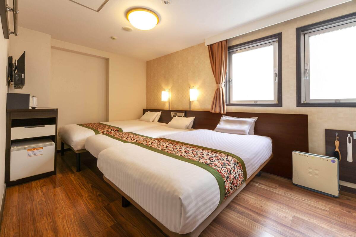 A hotel room with two beds, a desk, a minibar, and a window with curtains.
