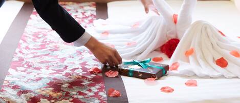 Eine Person richtet ein Bett mit einer floralen Tagesdecke, einer herzförmigen Schwan-Dekoration und einer Geschenkbox her.