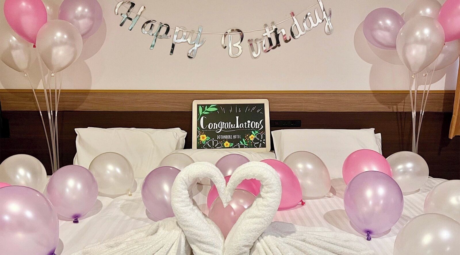 A hotel room with a bed, balloons, and a 'Happy Birthday' banner.