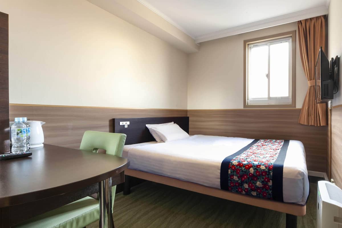 A hotel room with a single bed, a desk, a chair, a TV mounted on the wall, and a window with curtains.
