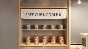 A shelf with multiple containers of instant noodles, a sign above stating "FREE CUP NOODLE," and a microwave in the background. - Dotonbori Hotel (Osaka)
