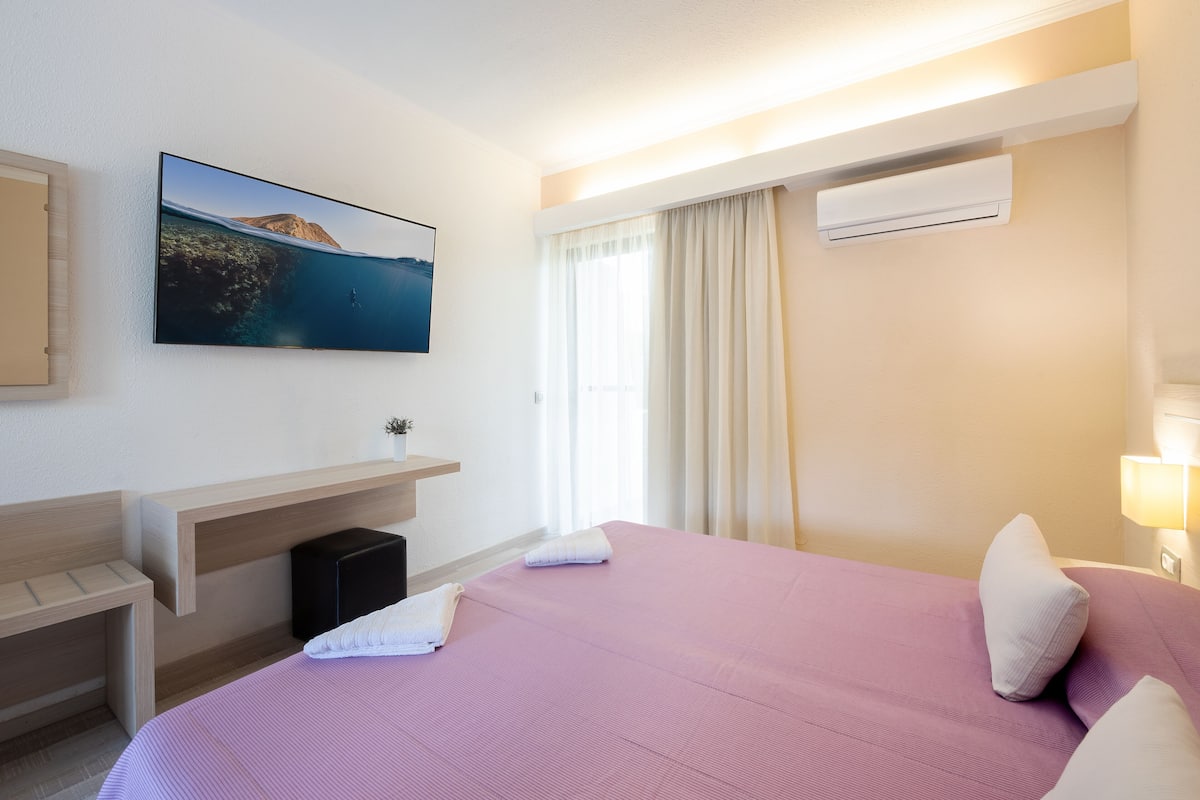 Una habitación de hotel con cama, televisor montado, escritorio y ventana con cortinas.