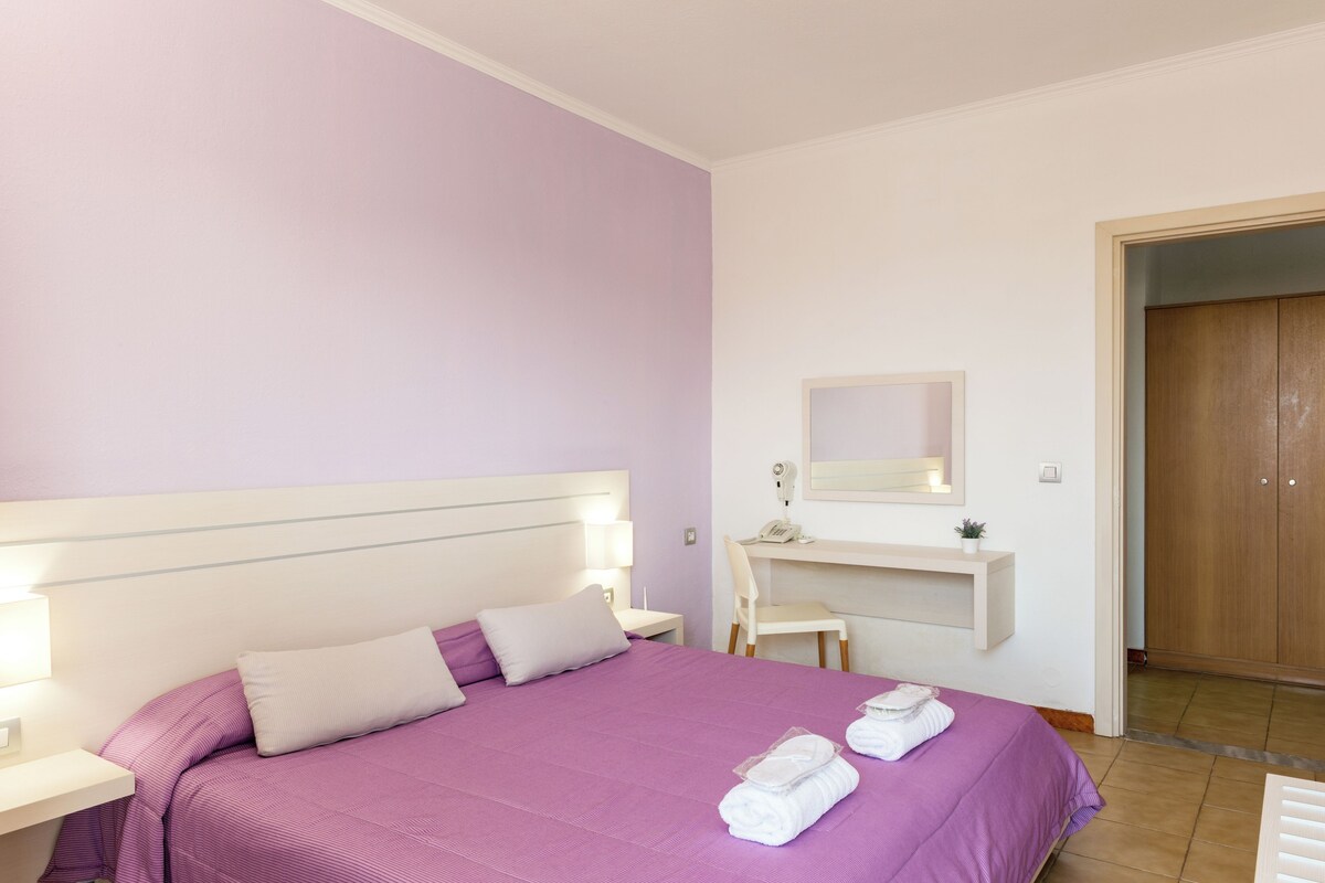 Quarto com cama, mesas de cabeceira, escrivaninha, cadeira e guarda-roupa.