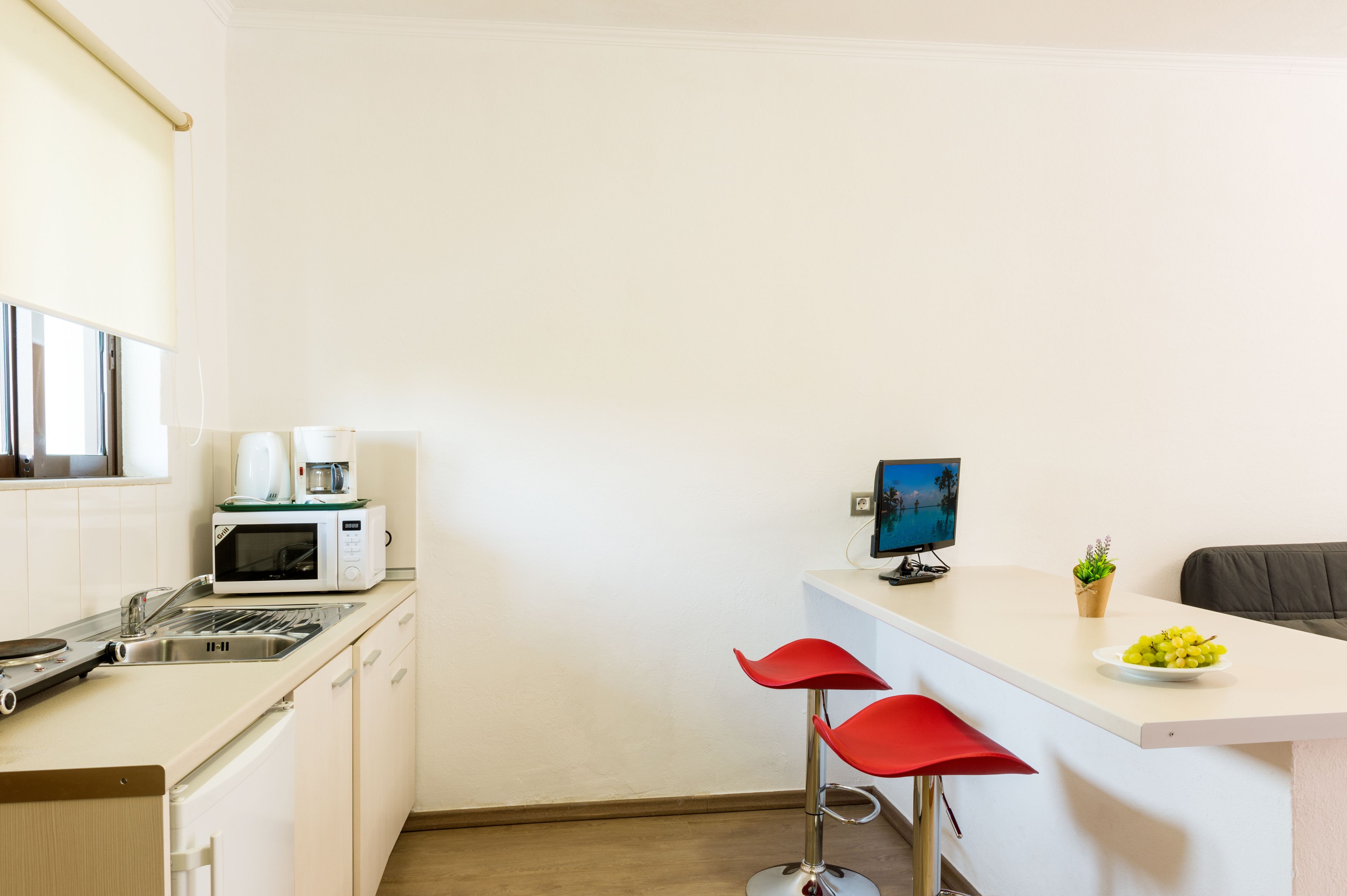 A compact kitchen with a microwave, coffee maker, and a small TV on a counter. There are two red bar stools and a potted plant on the counter.