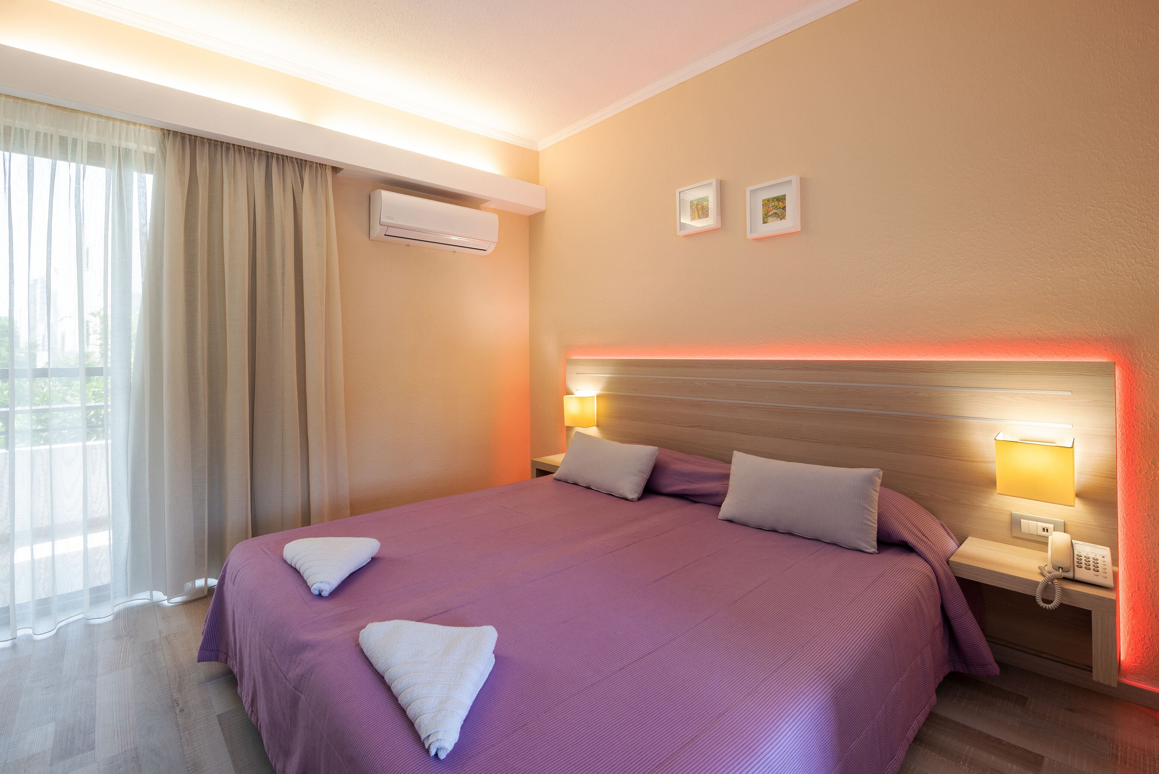 A hotel room with a large bed, two bedside tables with lamps, an air conditioning unit, and a window with curtains.