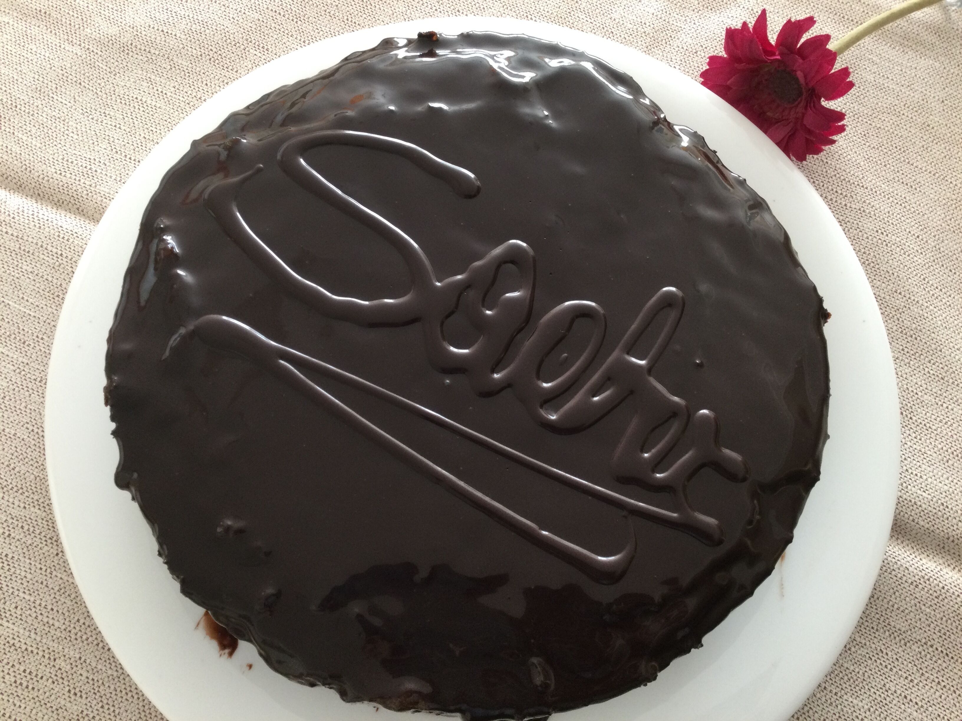 A chocolate-covered cake with the word "Soofer" written on it, placed on a white plate.