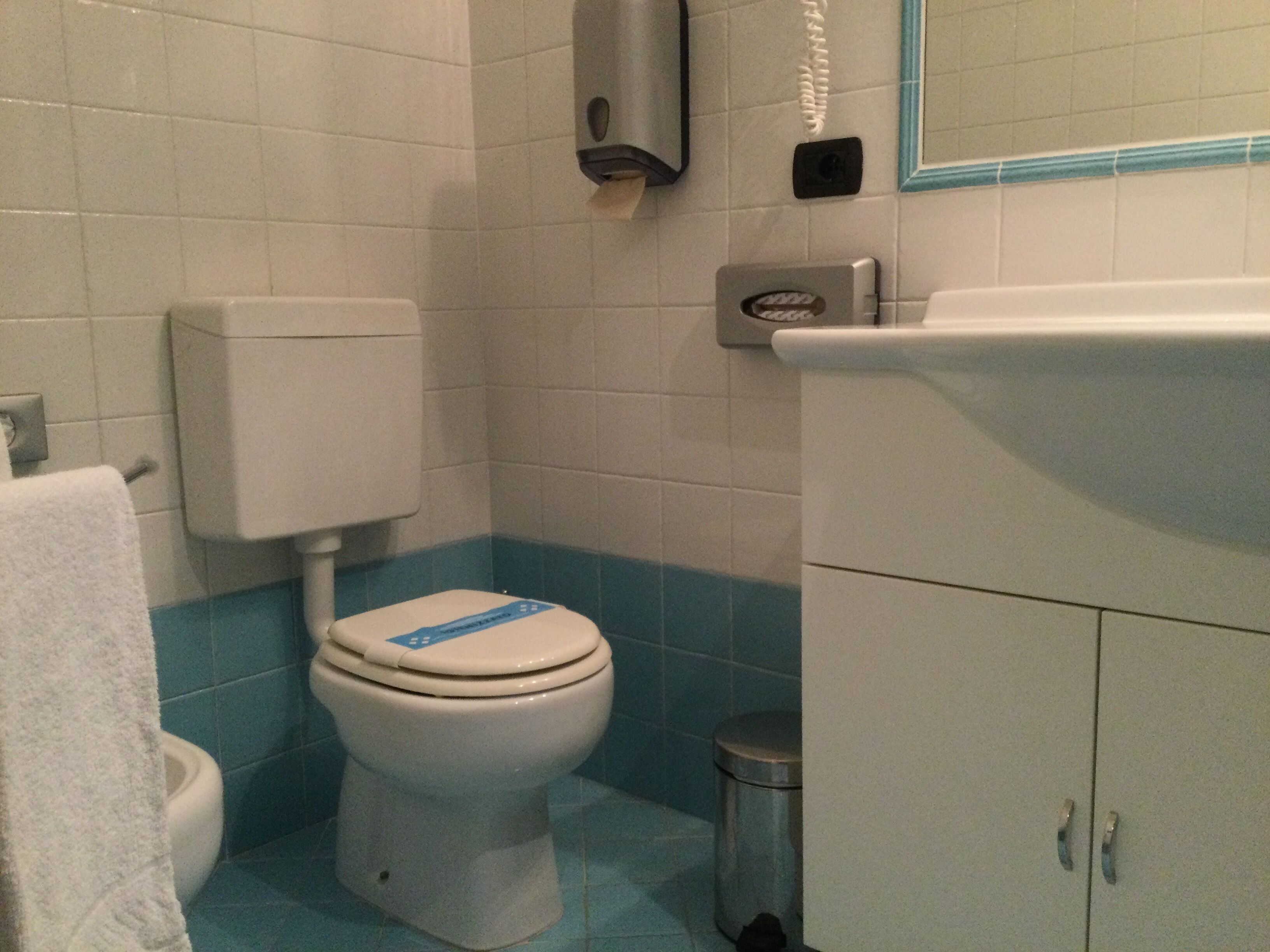 Un baño con inodoro, secamanos y cubo de basura.