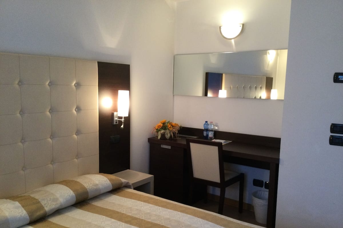 Habitación de hotel con cama, escritorio, silla, espejo y lámpara fijada a la pared.