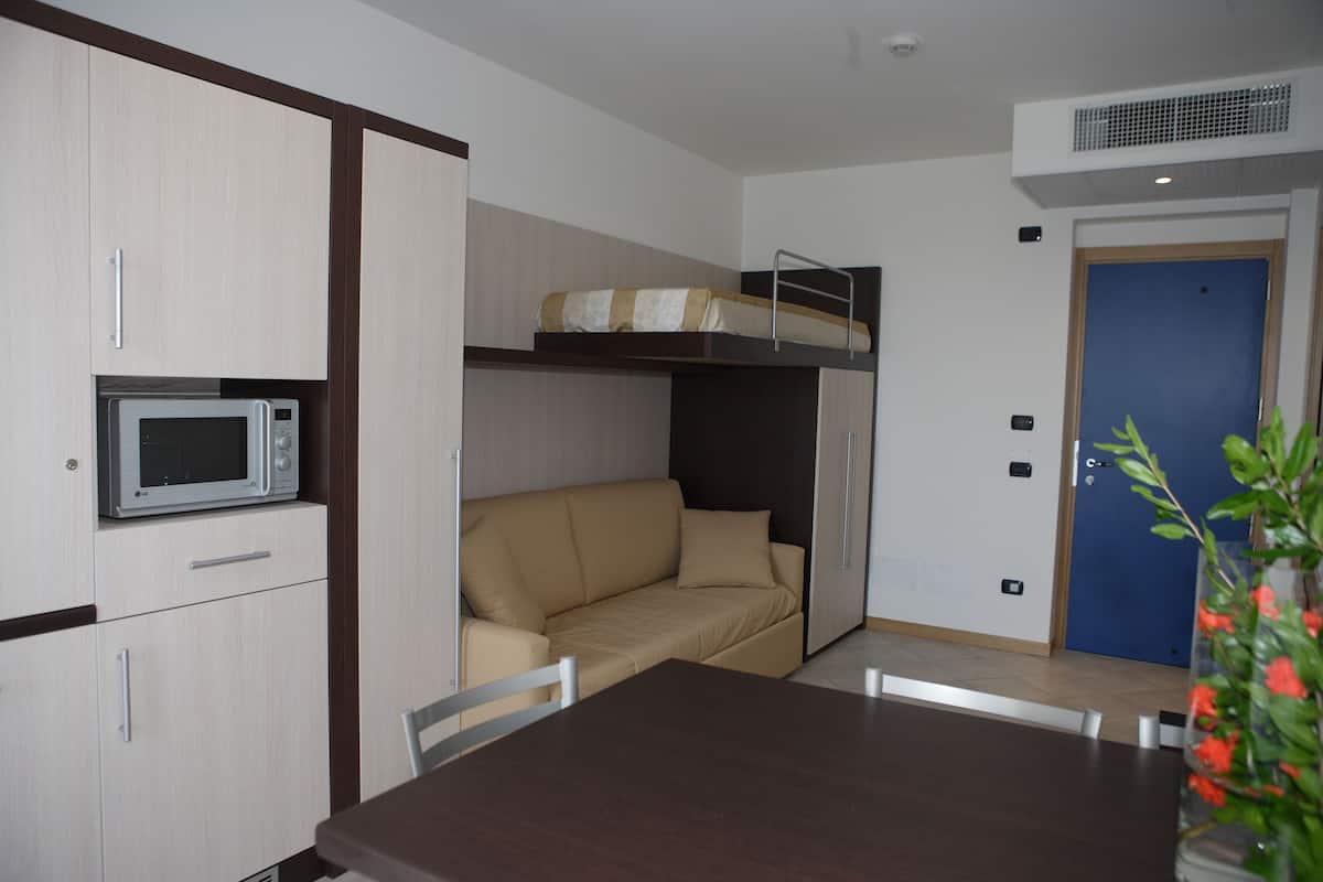 Una habitación compacta con una cama nido, un microondas, una mesa de comedor y un jarrón con flores.