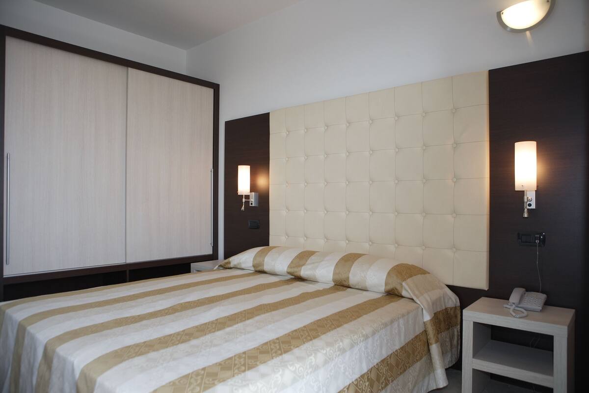 Una habitación de hotel con una cama grande, un armario, una mesita de noche con lámpara y un teléfono.