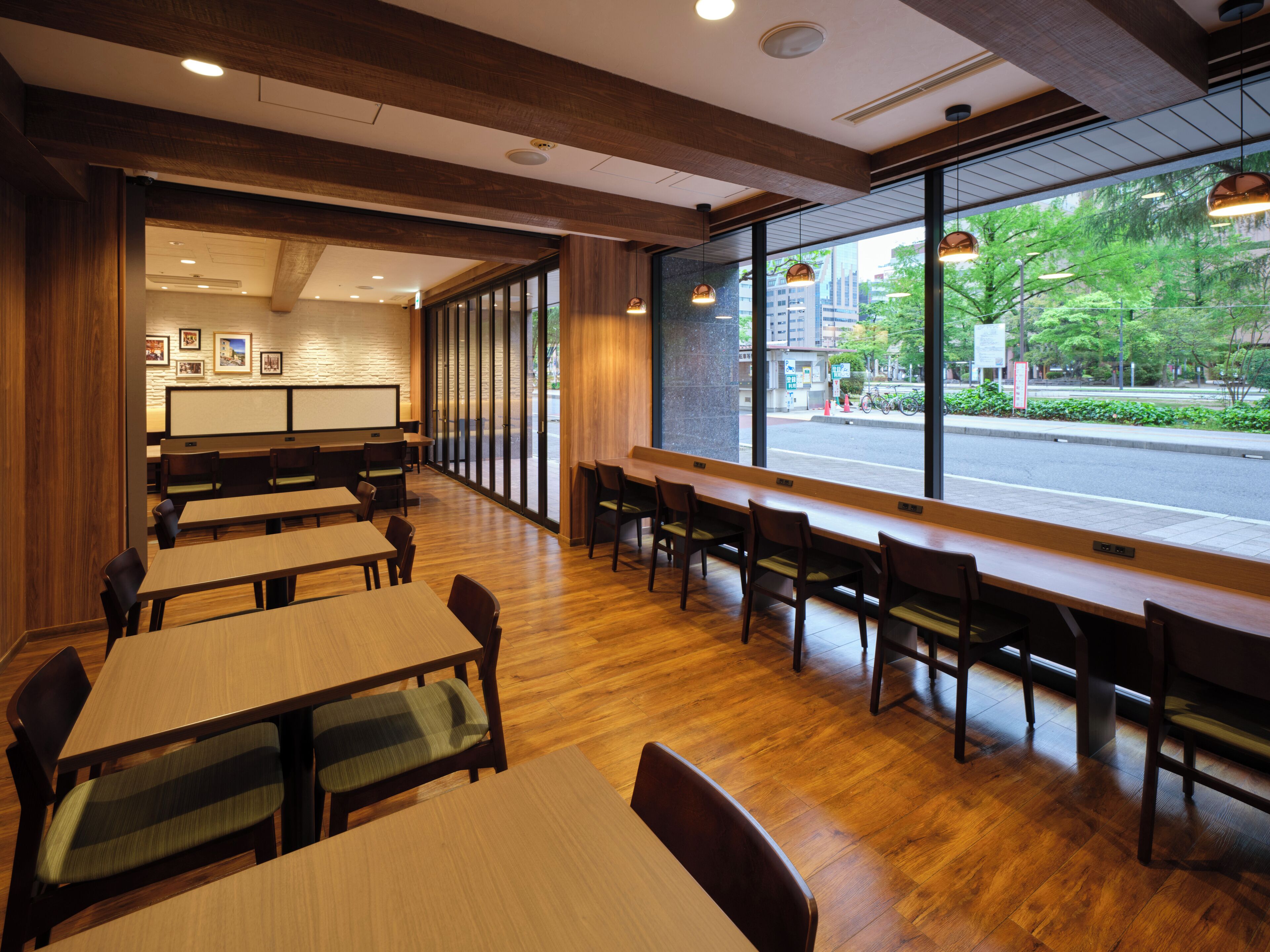 A restaurant interior with wooden tables and chairs, large windows offering a view of the street, and recessed ceiling lighting.