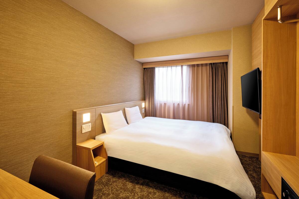 A hotel room with a large bed, a bedside table, a chair, a flat-screen TV, and a window with curtains.