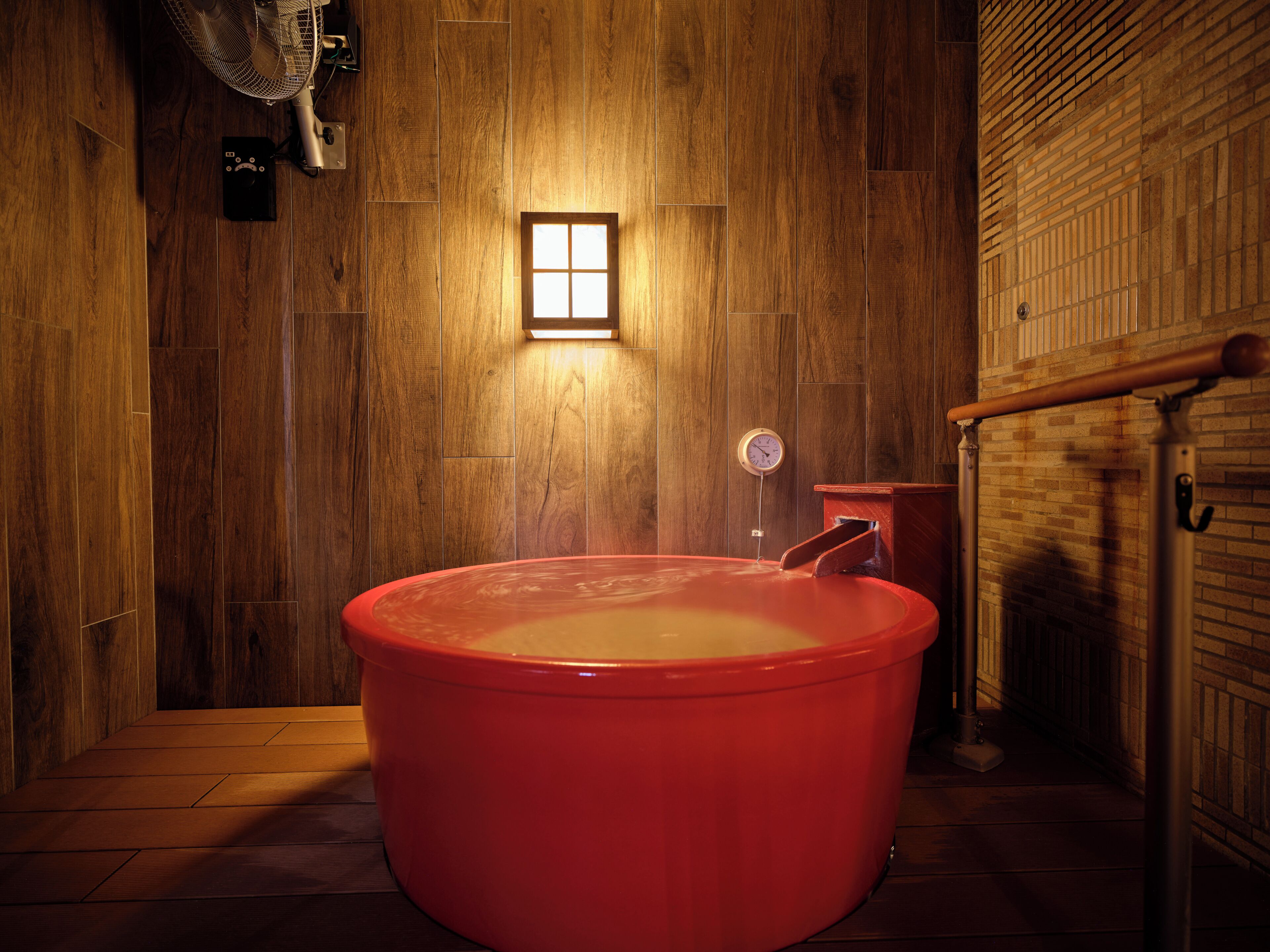 A wooden room with a large red bathtub, a small window, and a clock on the wall.