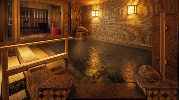 Un onsen japonés tradicional con interior de madera, una fuente termal de piedra y un banco.