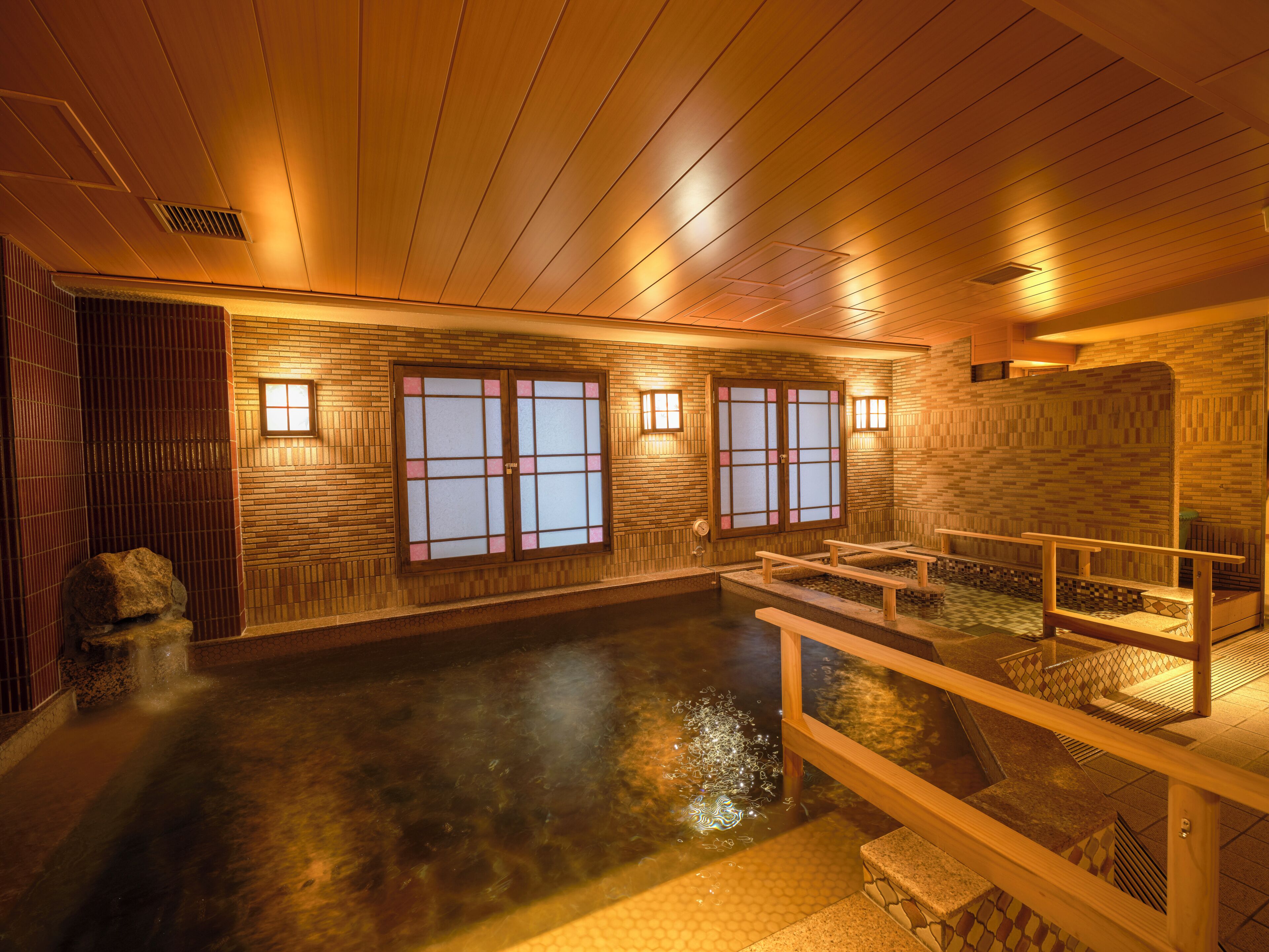 Un onsen japonés tradicional con suelos de madera, paredes de azulejos y techo de madera. Hay bancos a lo largo de la poza termal, y la habitación está iluminada por luces fijadas en la pared.