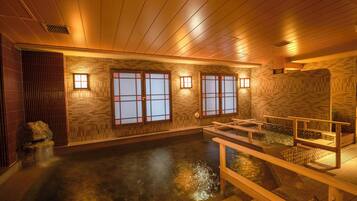 Un onsen japonés tradicional con suelos de madera, paredes de azulejos y techo de madera. Hay bancos a lo largo de la poza termal, y la habitación está iluminada por luces fijadas en la pared.
