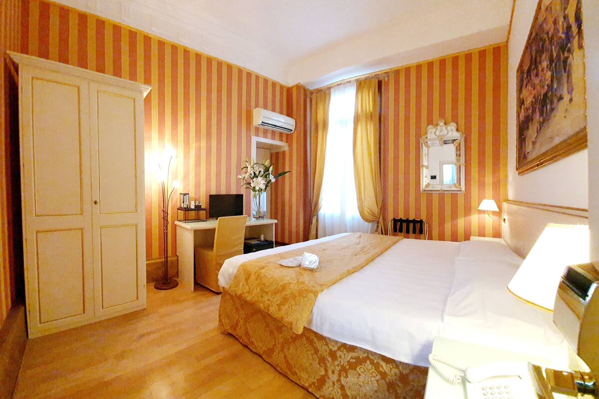 Habitación de hotel con cama, mesita de noche, escritorio, silla, armario y equipo de aire acondicionado.