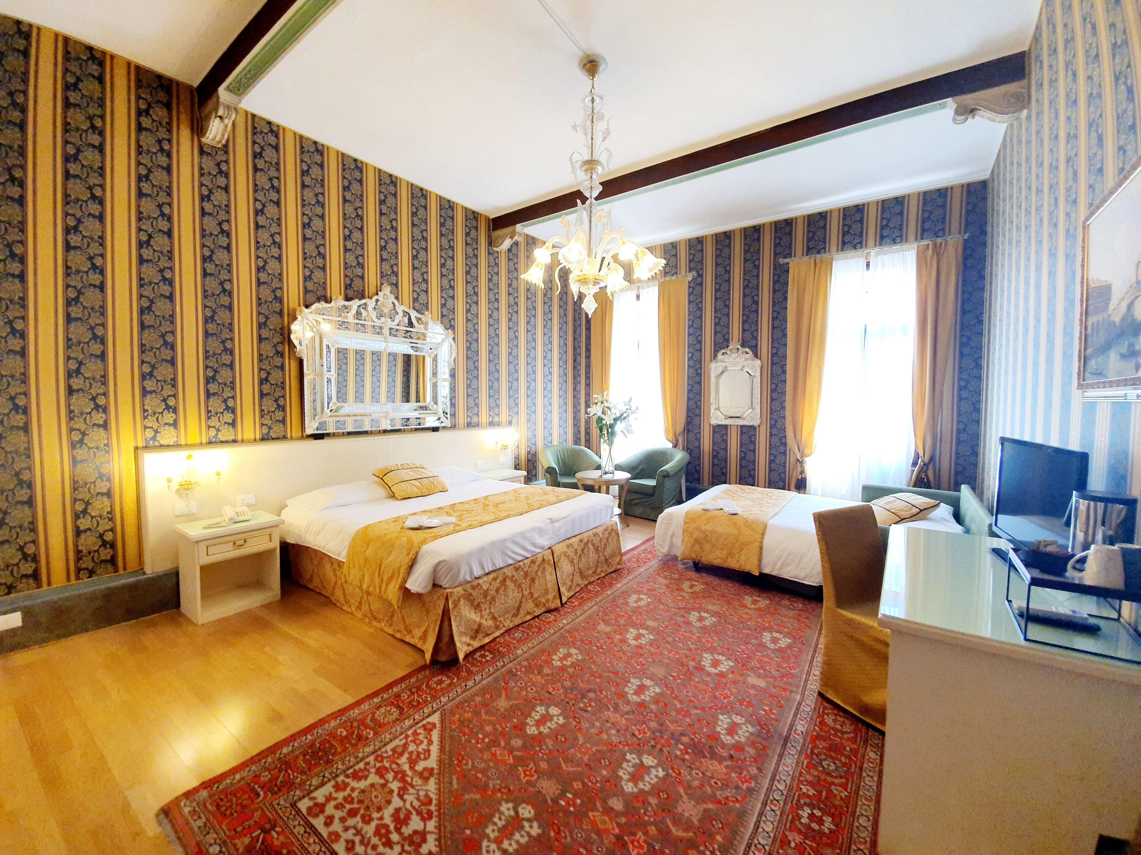 A hotel room with two beds, a large mirror, a chandelier, a desk with a computer, and a TV.