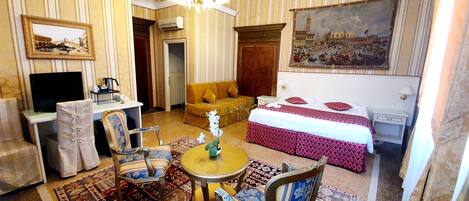 A hotel room with a bed, a sofa, a desk with a computer, a television, and a chandelier.