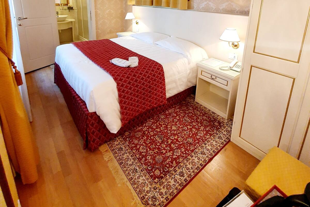 Habitación de hotel con una cama, mesitas de noche, un espejo, una ventana con cortinas, una alfombra y una puerta.