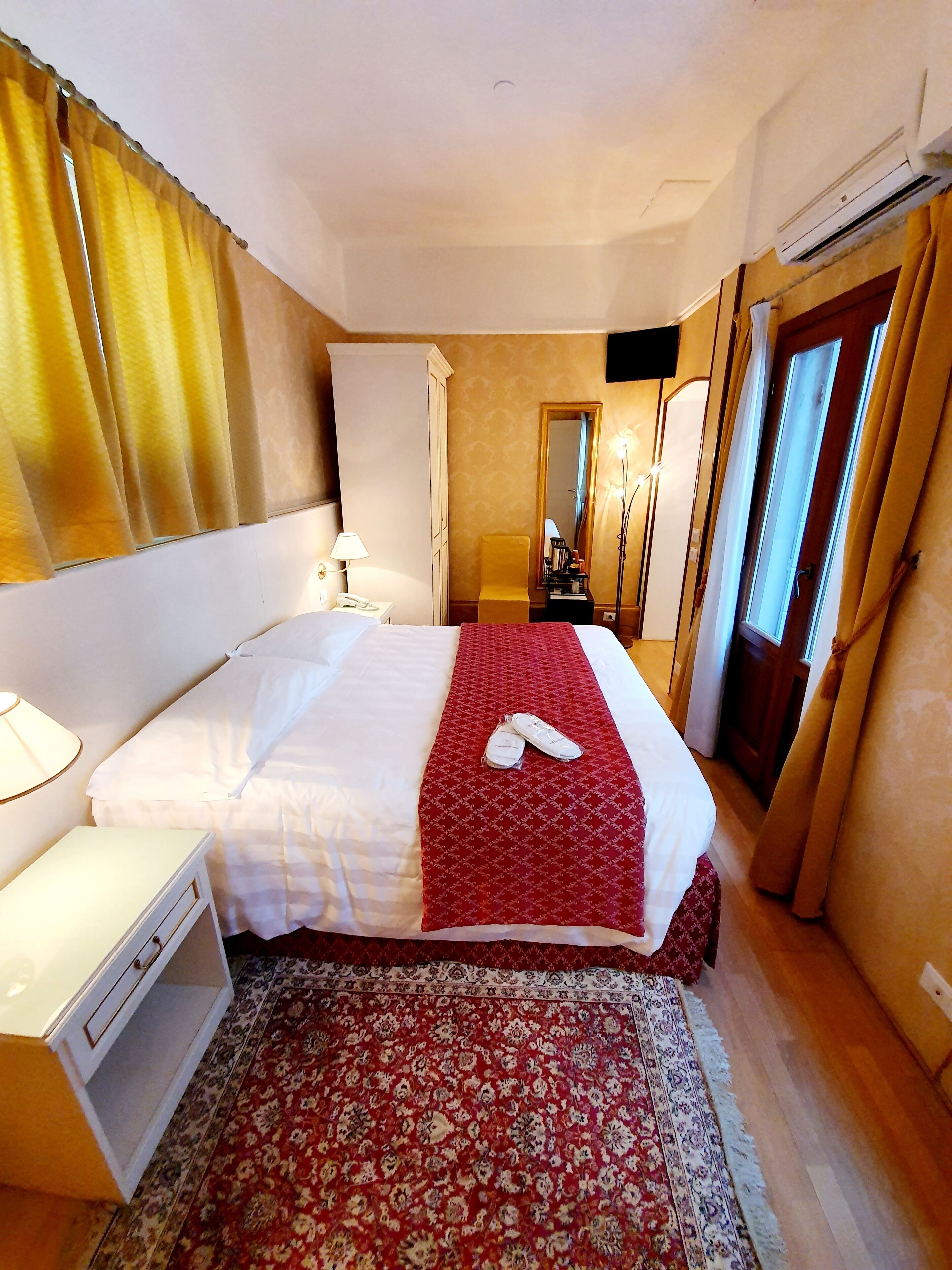 A hotel room with a bed, bedside table, lamp, and a red patterned rug.