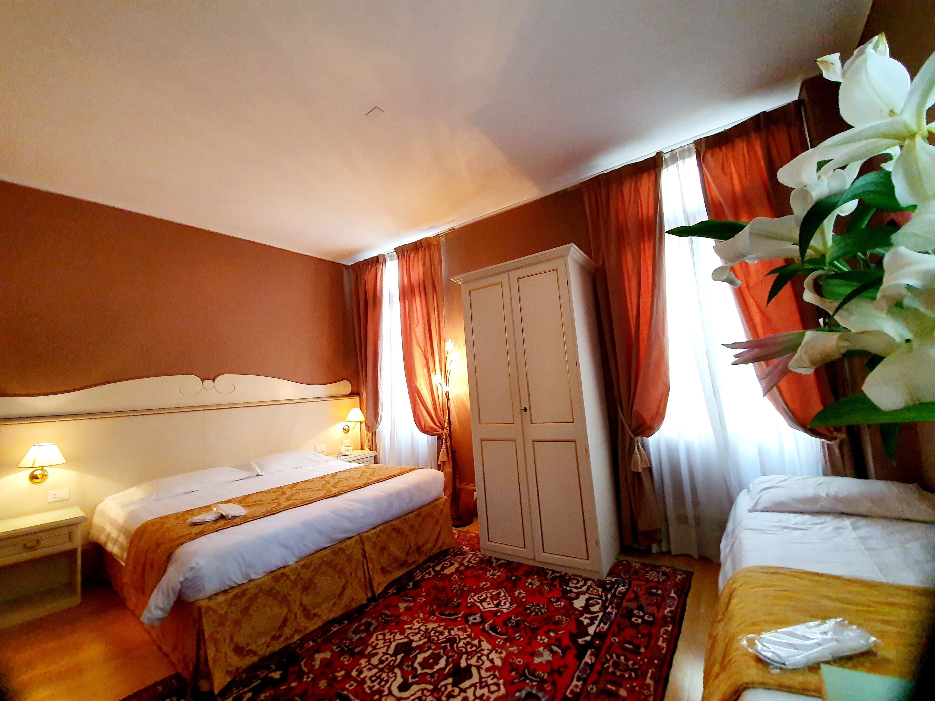 A hotel room with two beds, a wardrobe, a red patterned rug, and a large white flower.