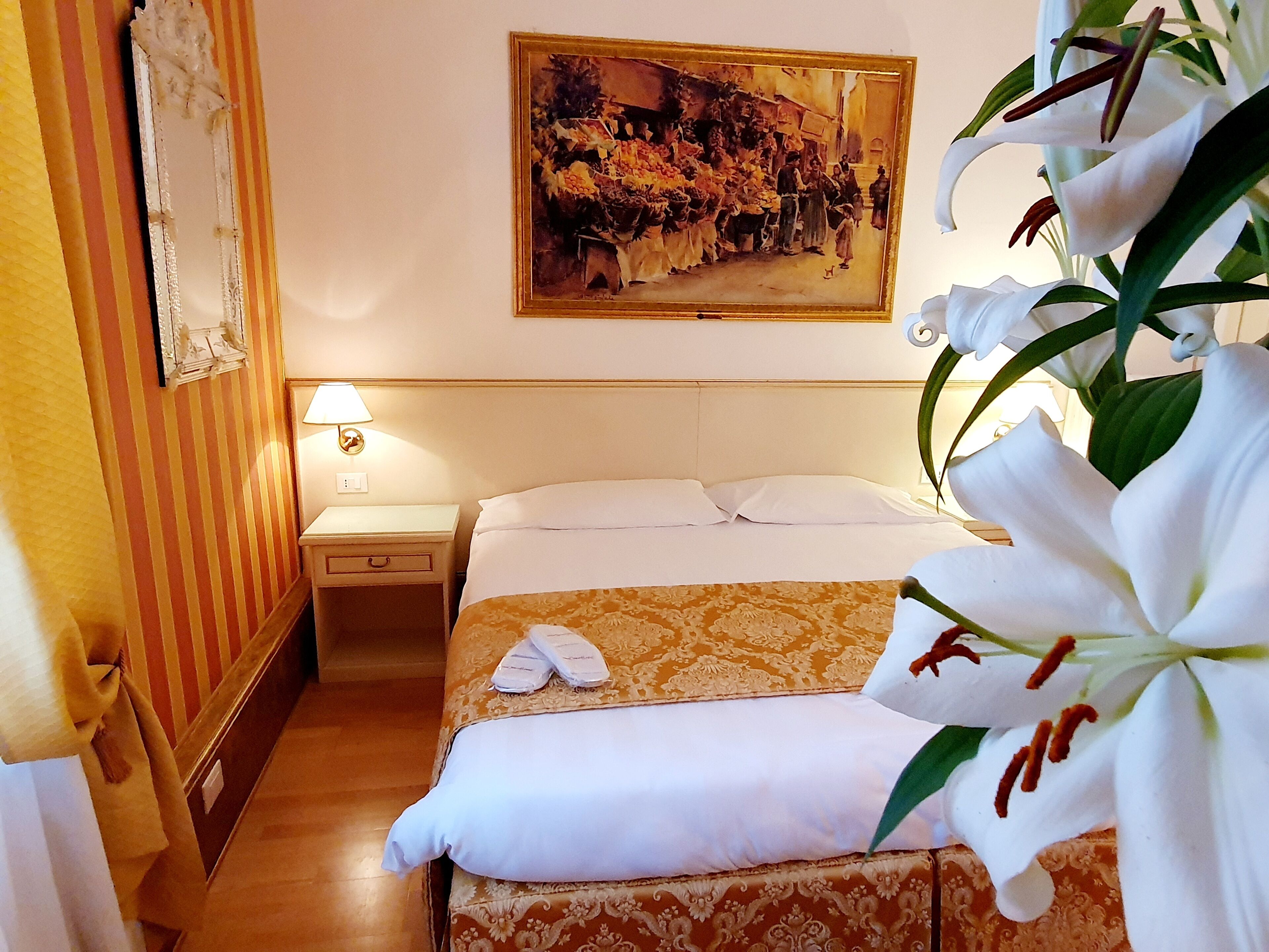 A hotel room with a bed, a nightstand, a lamp, a painting on the wall, and a potted plant.
