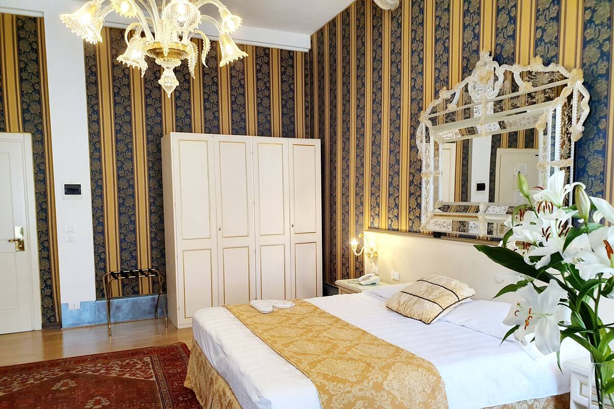 Habitación de hotel con una cama grande, un candelabro, un armario y un espejo con motivos florales.