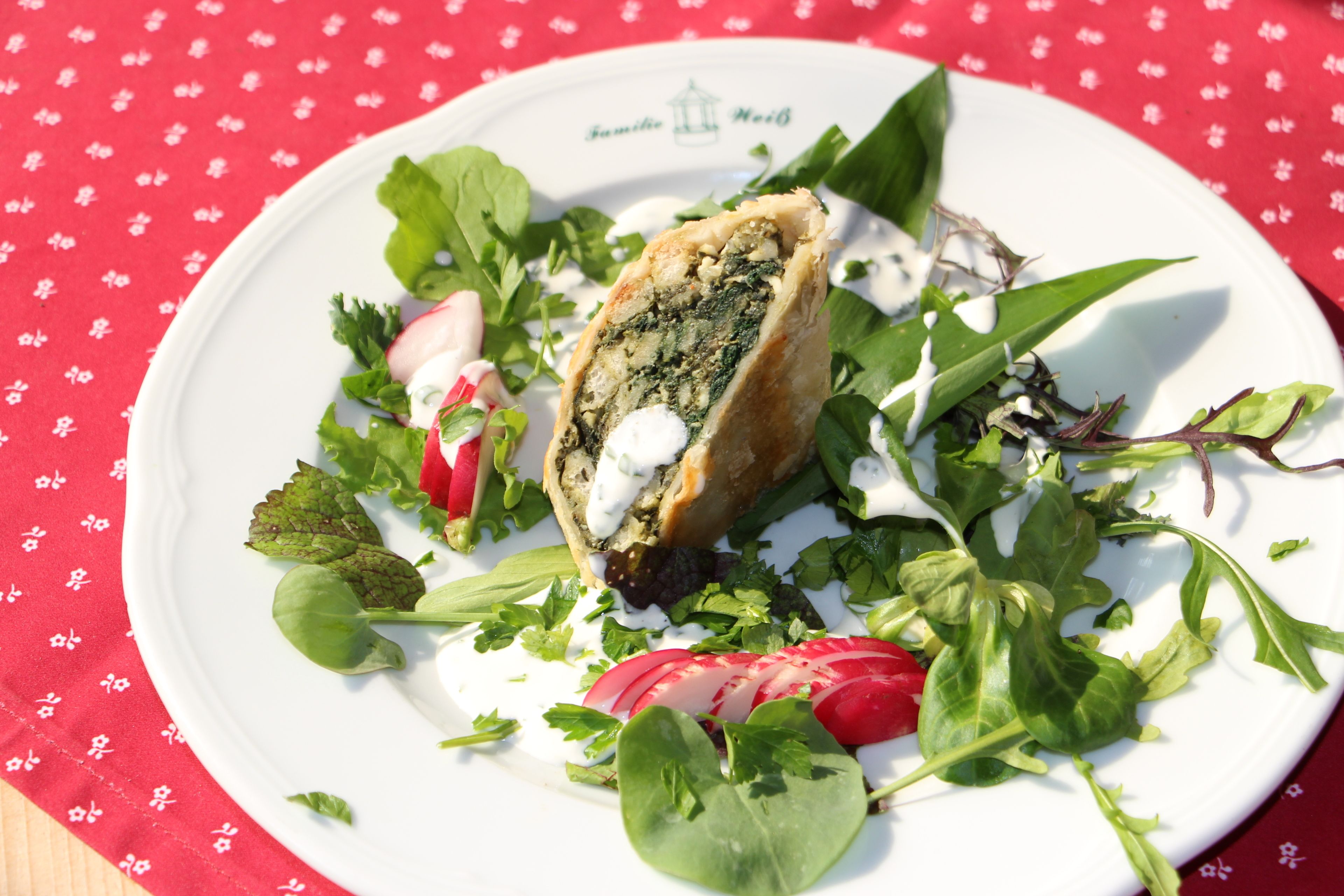 A plate with a pastry, greens, and radishes on a tablecloth.
