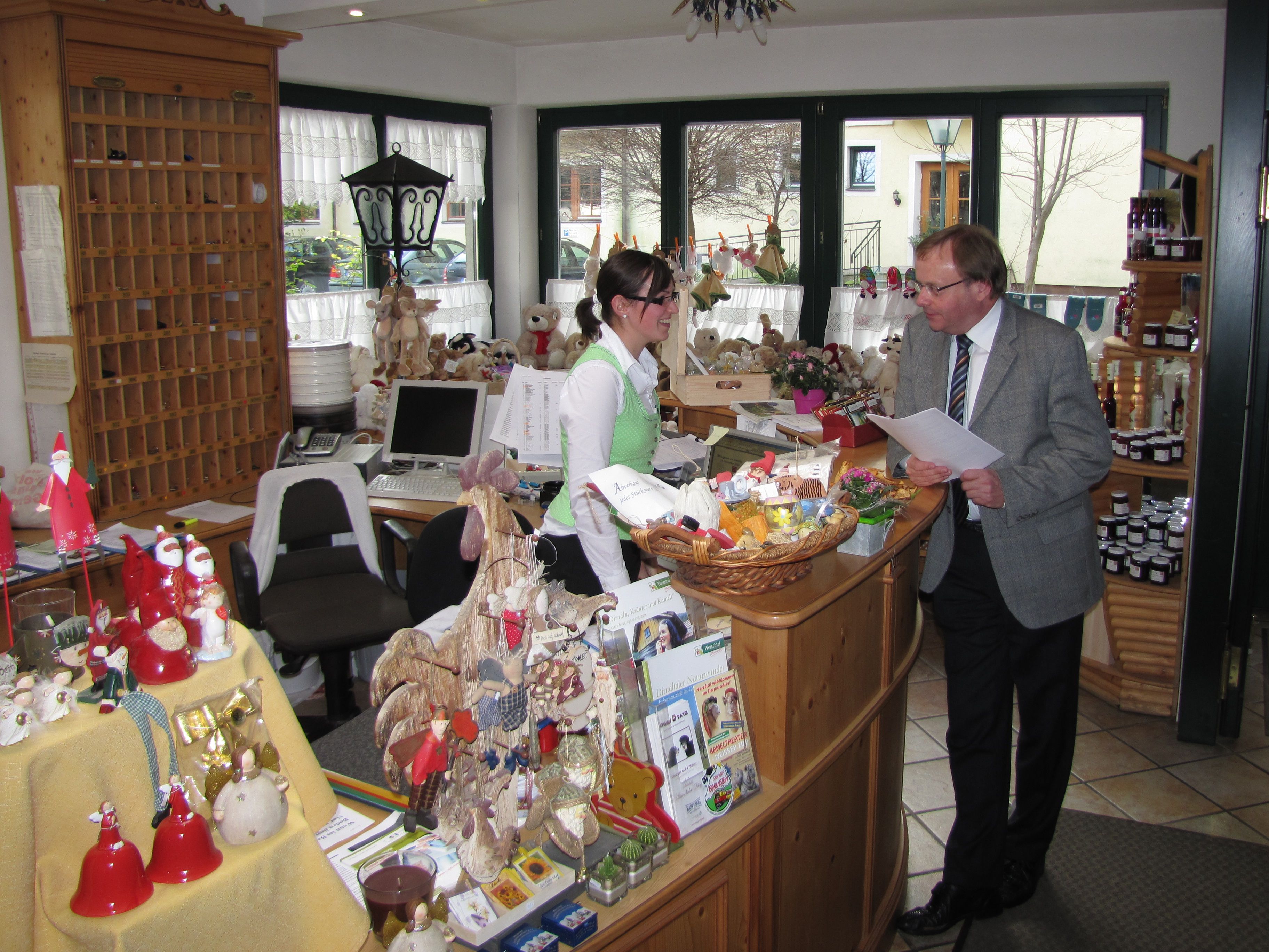 A shop with a wooden counter displaying various items, a computer, and a window with a view of the outside.