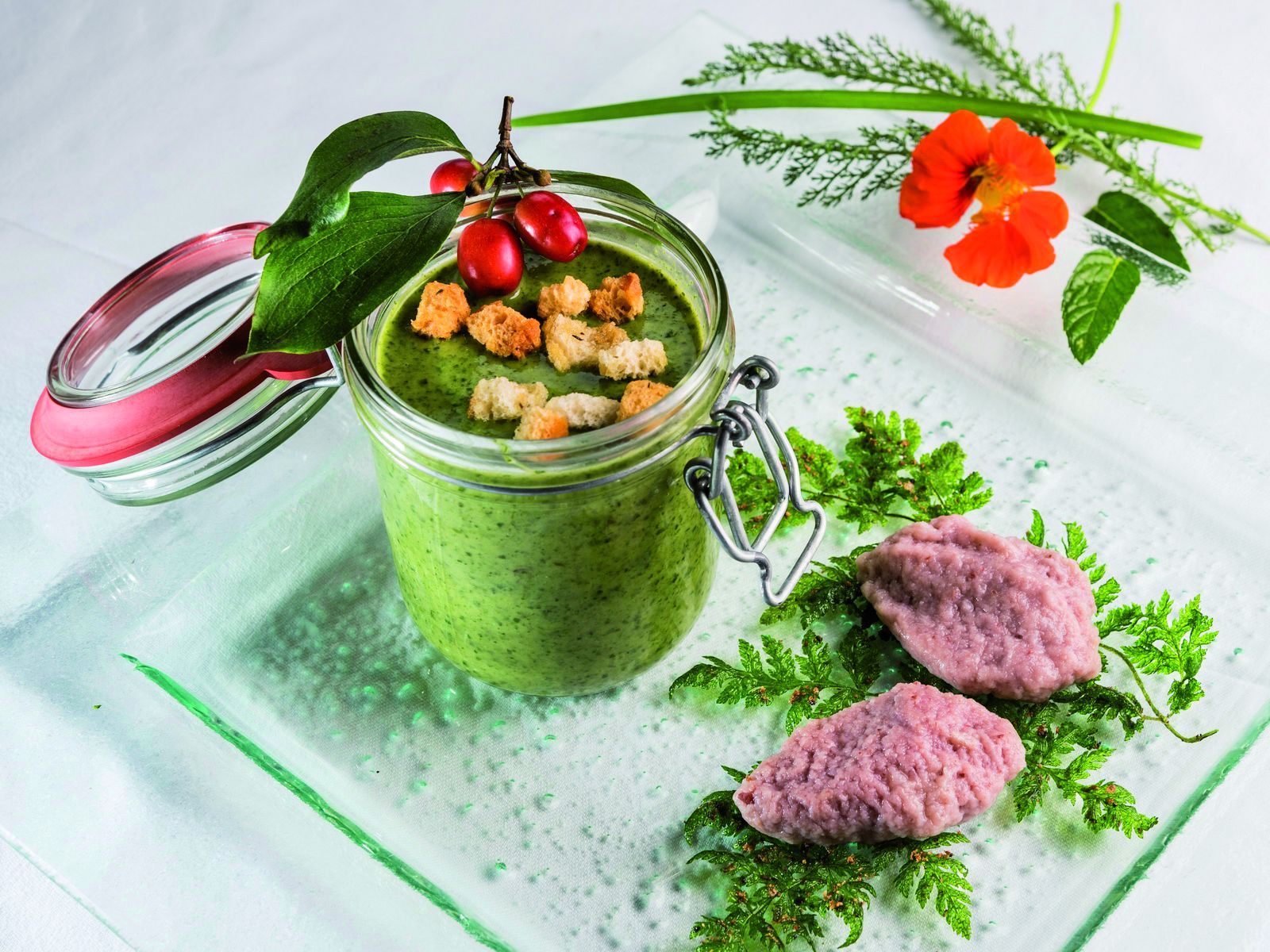 Um frasco de smoothie verde com croutons e frutas vermelhas, decorado com salsa e uma flor vermelha.