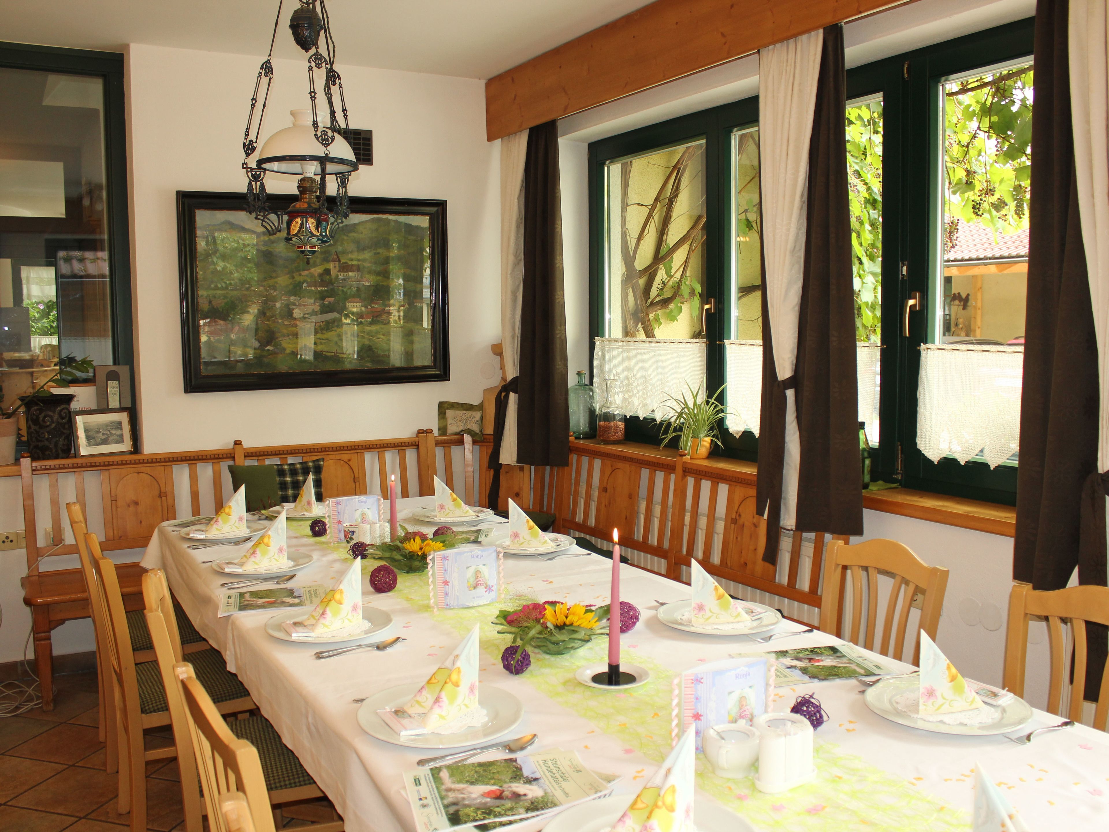 Un comedor con una mesa larga dispuesta para una comida, sillas de madera, una araña y una ventana con cortinas.