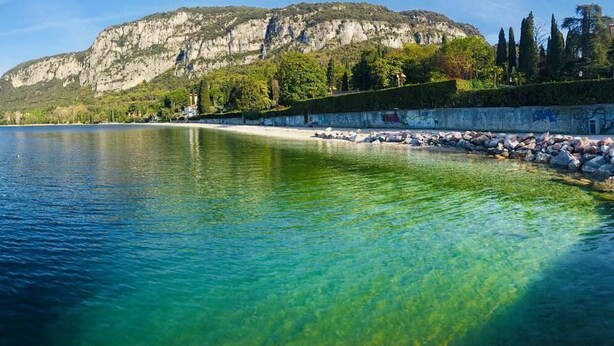 Un lago transparente con costa rocosa y una montaña al fondo.