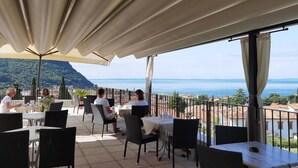 Un área de comedor al aire libre con mesas y sillas, vista al mar y montañas de fondo.