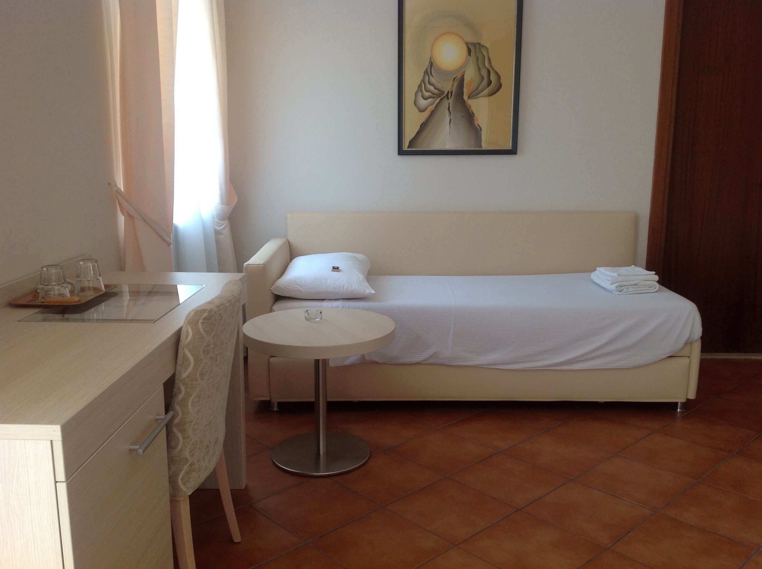 Quarto de hotel com cama, mesa redonda pequena, escrivaninha e cadeira, e um quadro na parede.