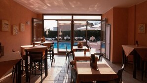 Un comedor interior con mesas y sillas, ventanas amplias y vistas a una piscina y sombrillas afuera.