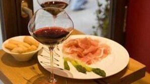 Una mesa de comedor con un plato de comida, una copa de vino tinto y un bol de palitos de pan.
