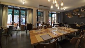 Un restaurante con mesas y sillas de madera, lámparas colgantes y grandes ventanales con cortinas.