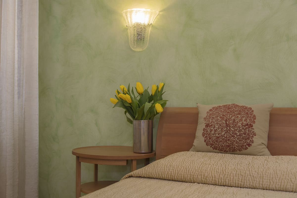 Ein Schlafzimmer mit grüner Wand, einem Nachttisch mit einer Vase gelber Blumen, einem hölzernen Kopfteil und einem Zierkissen.