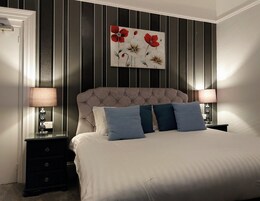 Quarto com cabeceira estofada, cama com roupa de cama branca e almofadas azuis, duas mesas de cabeceira com abajures e um quadro de flores na parede.