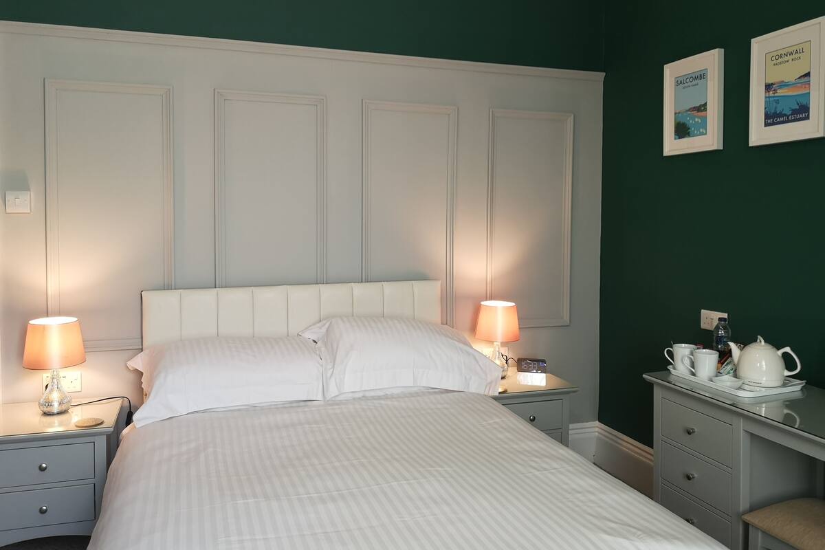 A neatly made bed with white linens, two bedside lamps, a nightstand with a clock, a small table with a teapot and cups, and two framed pictures on the wall.