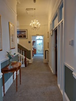 A hallway with a chandelier, framed pictures, a vase with flowers, and a staircase.